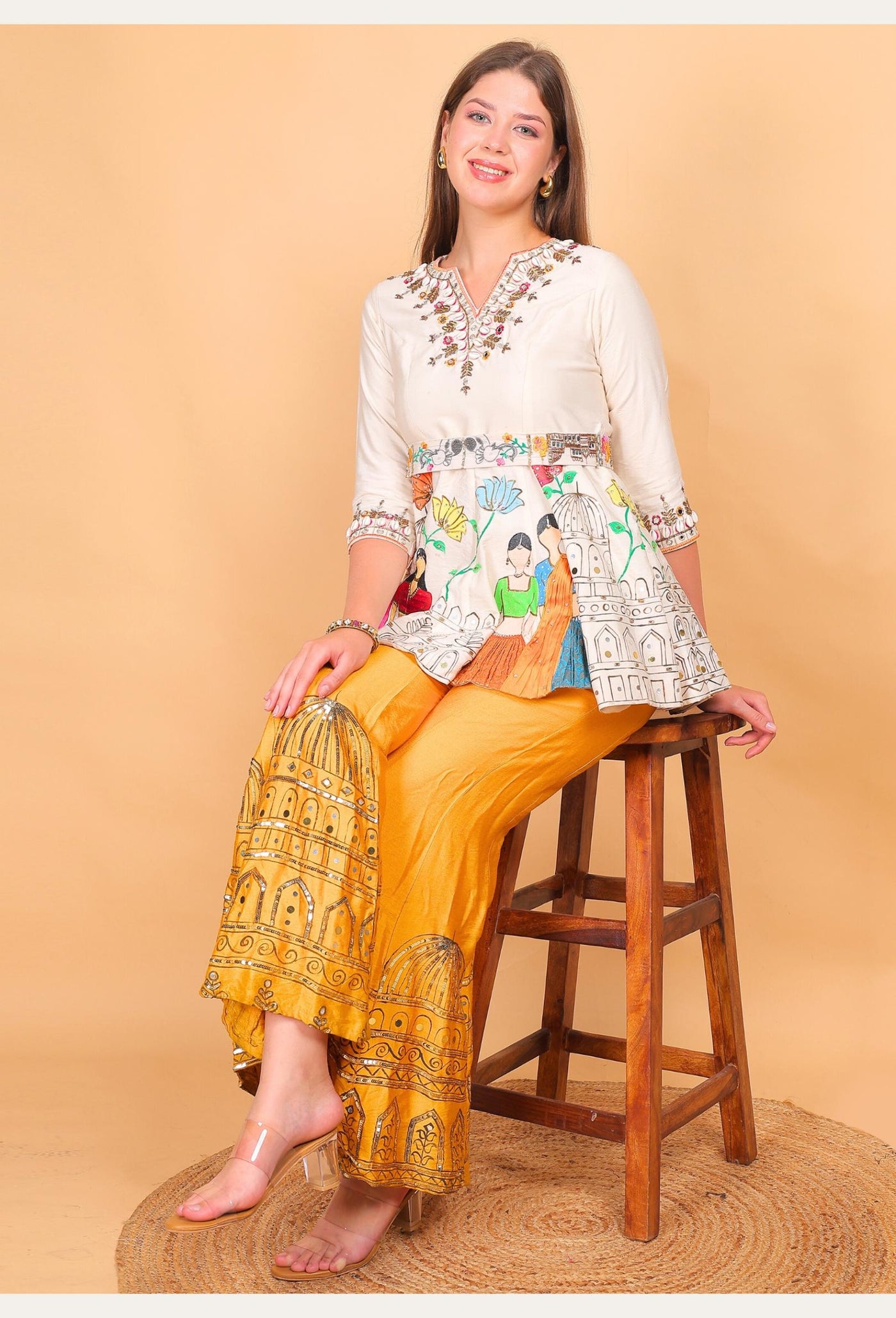 Woman sitting on a wooden stool wearing a patterned top and yellow pants against a beige background